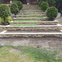 Kamienne schody tarasowe prowadzące do domu, z rabatami obsadzonymi trawą i kulistymi tujami, widok z dołu na dom z balkonem.