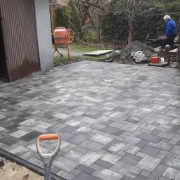 Ułożona kostka brukowa w szaro-czarnych odcieniach z widoczną łopatą na pierwszym planie, w tle betoniarka i osoba pracująca na posesji.