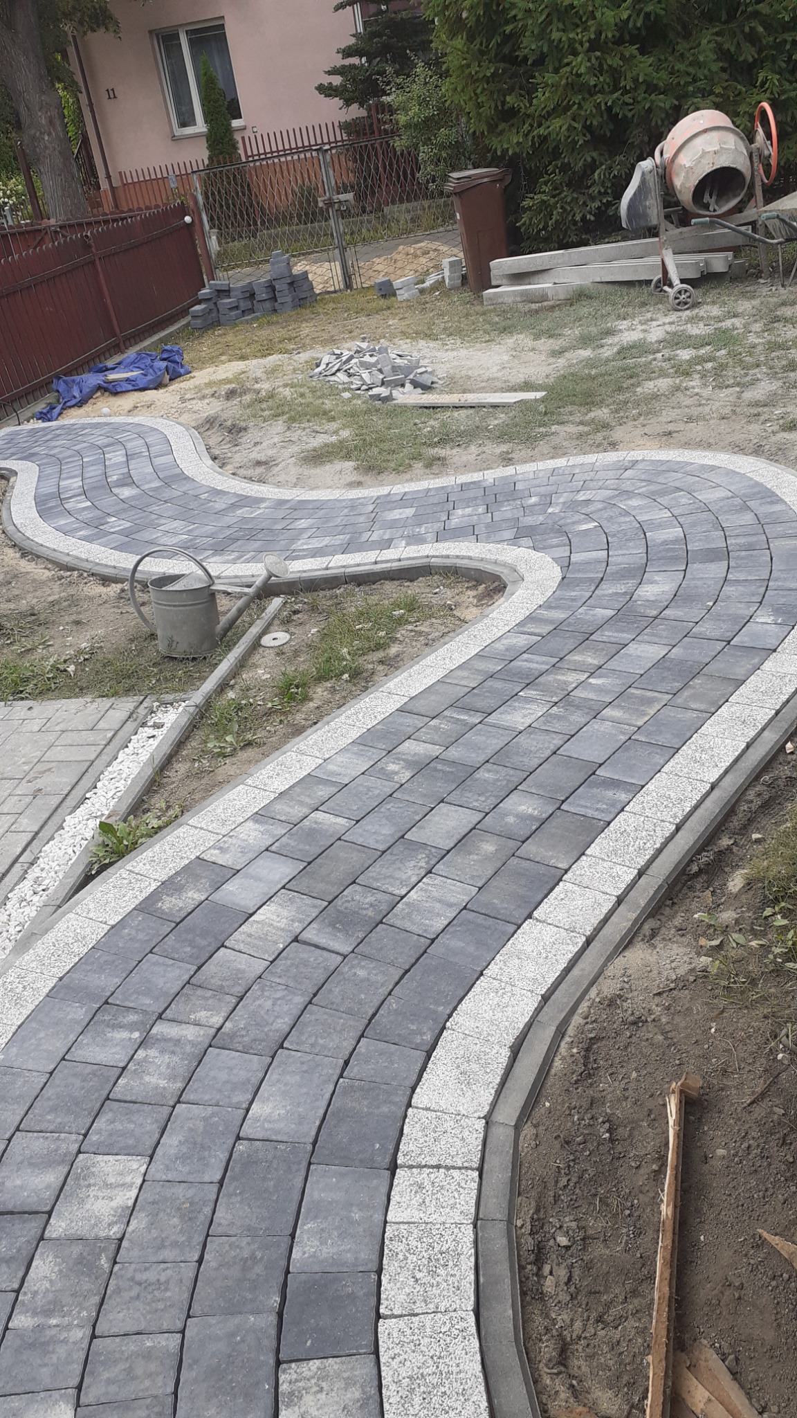 Kręta ścieżka z szarej kostki brukowej z jasnoszarym obramowaniem, widoczna z perspektywy ziemi, z konewką i mieszarką do betonu w tle.