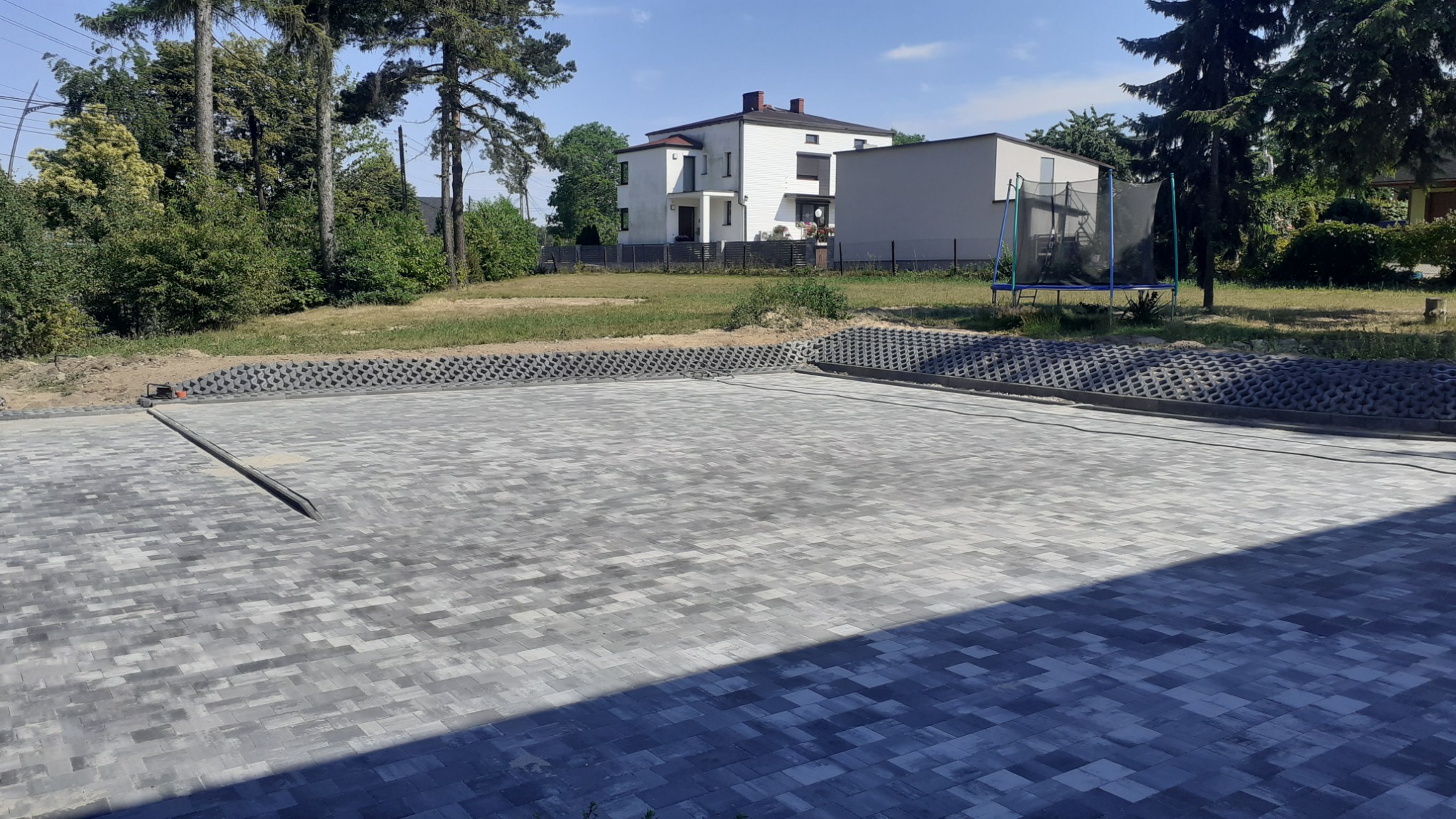 Ułożona szara kostka brukowa w jodełkę na posesji z domem i trampoliną w tle, widoczna linia odpływowa i ażurowe obrzeża.