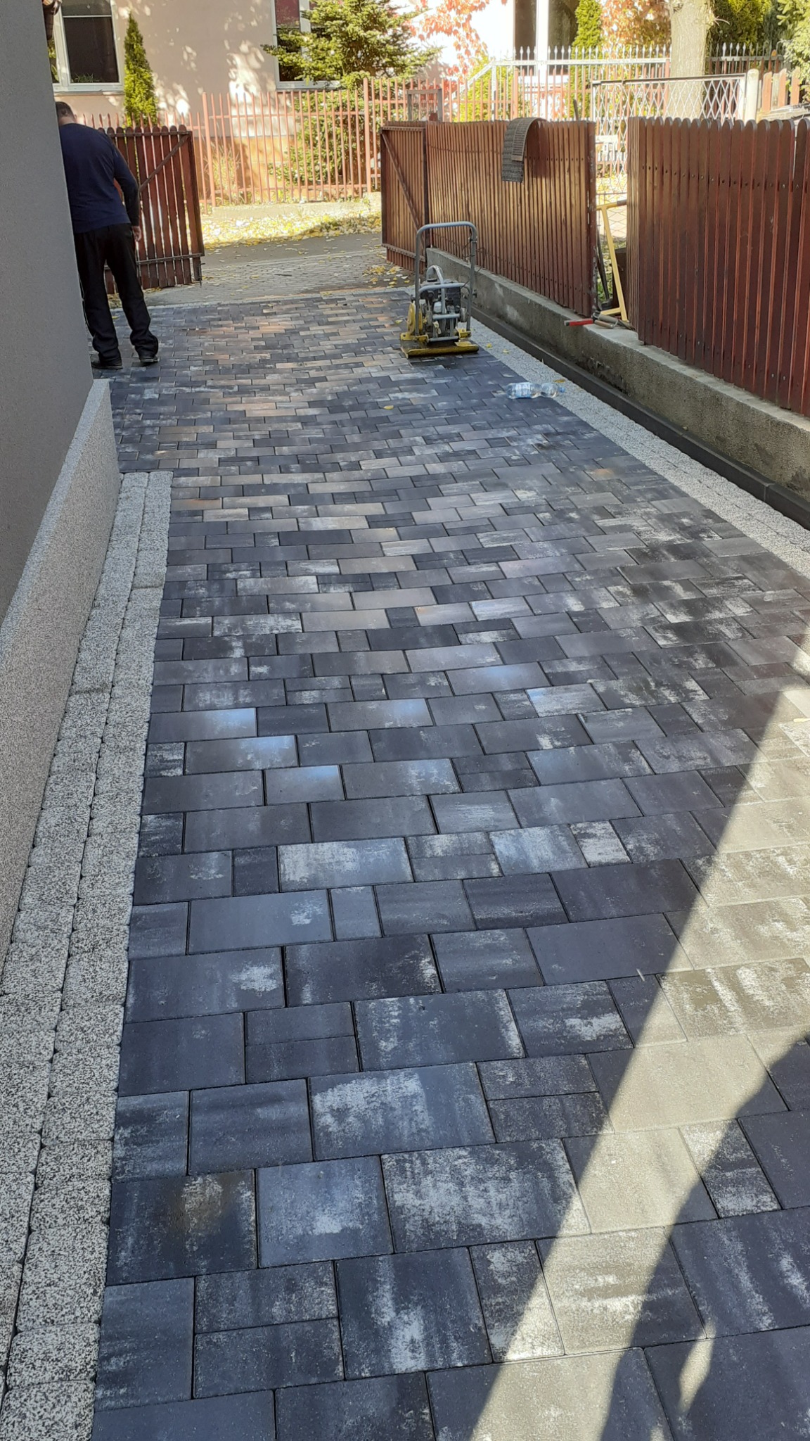 Świeżo ułożona, prostokątna kostka brukowa w odcieniach szarości z delikatnym wzorem, obrzeża z jasnego grysu, ubijarka w tle, częściowo widoczna osoba, brązowe ogrodzenie.