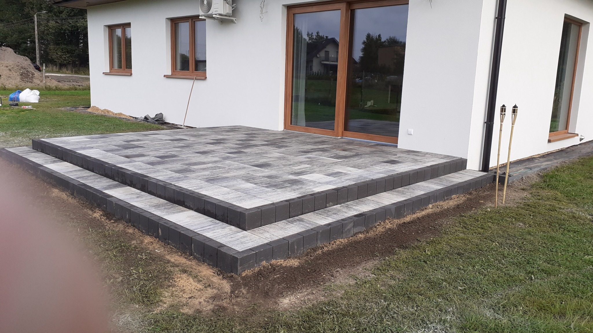 Nowoczesny taras z kostki brukowej w odcieniach szarości, prowadzący do domu z przesuwnymi drzwiami tarasowymi, z dwoma bambusowymi pochodniami po prawej stronie.