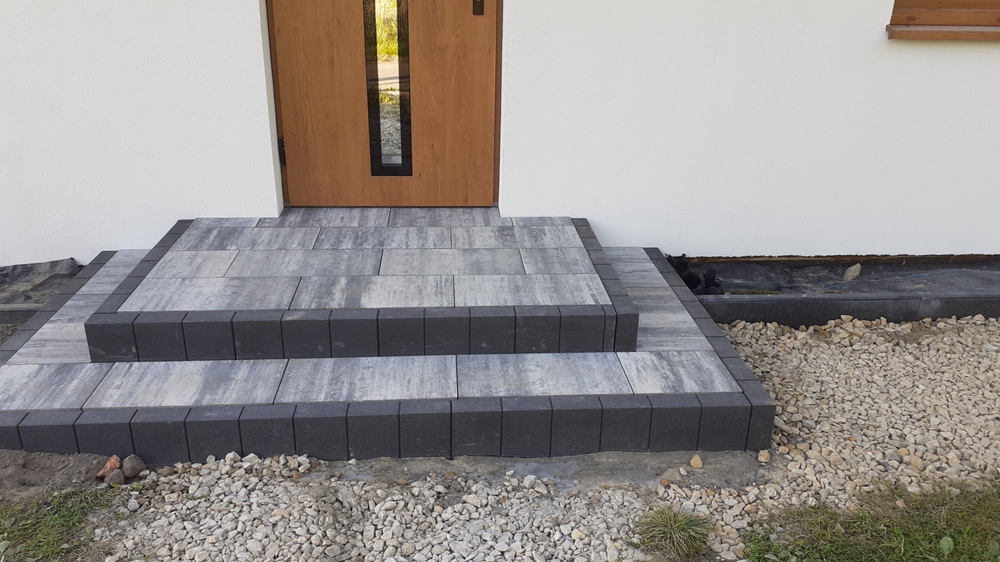 Schody wejściowe do domu wyłożone szarą kostką brukową z ciemnoszarą obwódką, widok z poziomu gruntu, drzwi wejściowe w tle.