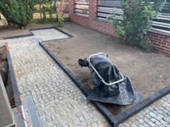 Fragment posesji w trakcie układania nawierzchni z kostki granitowej. Widoczna ułożona ścieżka z jasnej kostki granitowej obramowana ciemnym obrzeżem, oraz przygotowany teren pod dalsze prace.