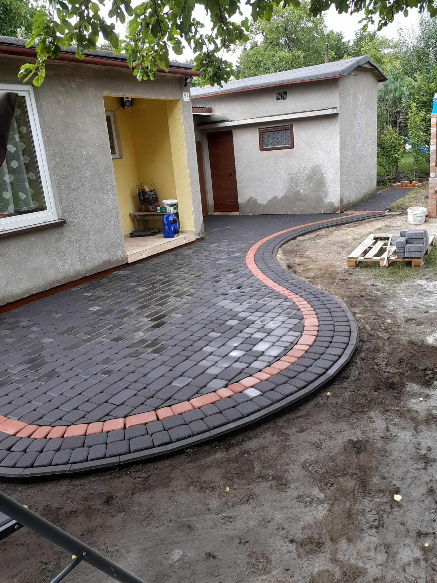Świeżo ułożony, zakrzywiony chodnik z ciemnoszarej kostki brukowej z obramowaniem z czerwonej kostki, prowadzący do budynku z żółtym podcieniem. Na palecie obok leżą kostki brukowe.