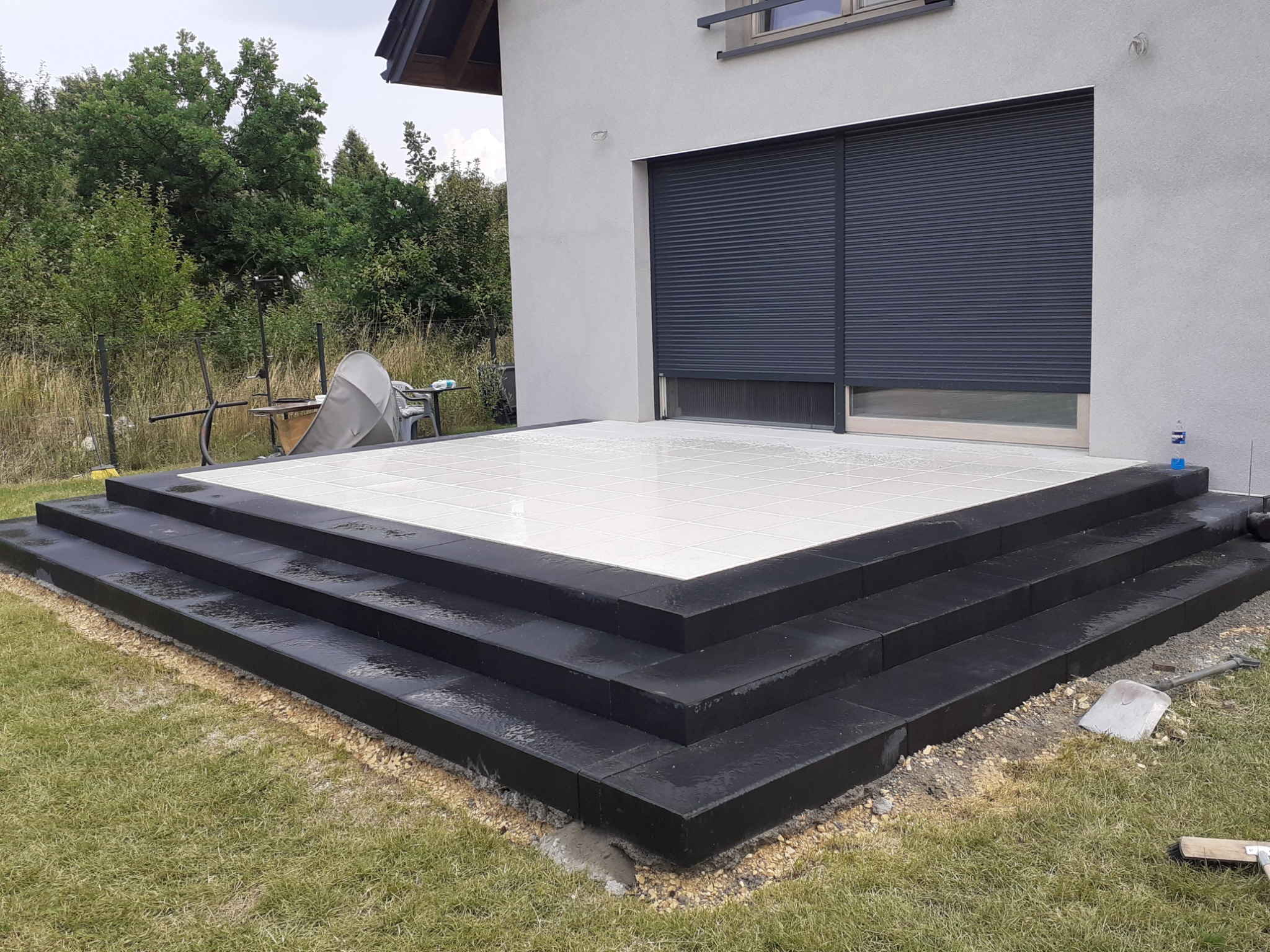 Nowoczesny taras z białych płytek, obramowany czarną kostką, prowadzący do budynku z szarymi roletami zewnętrznymi; widoczna łopata i szczotka na trawniku.