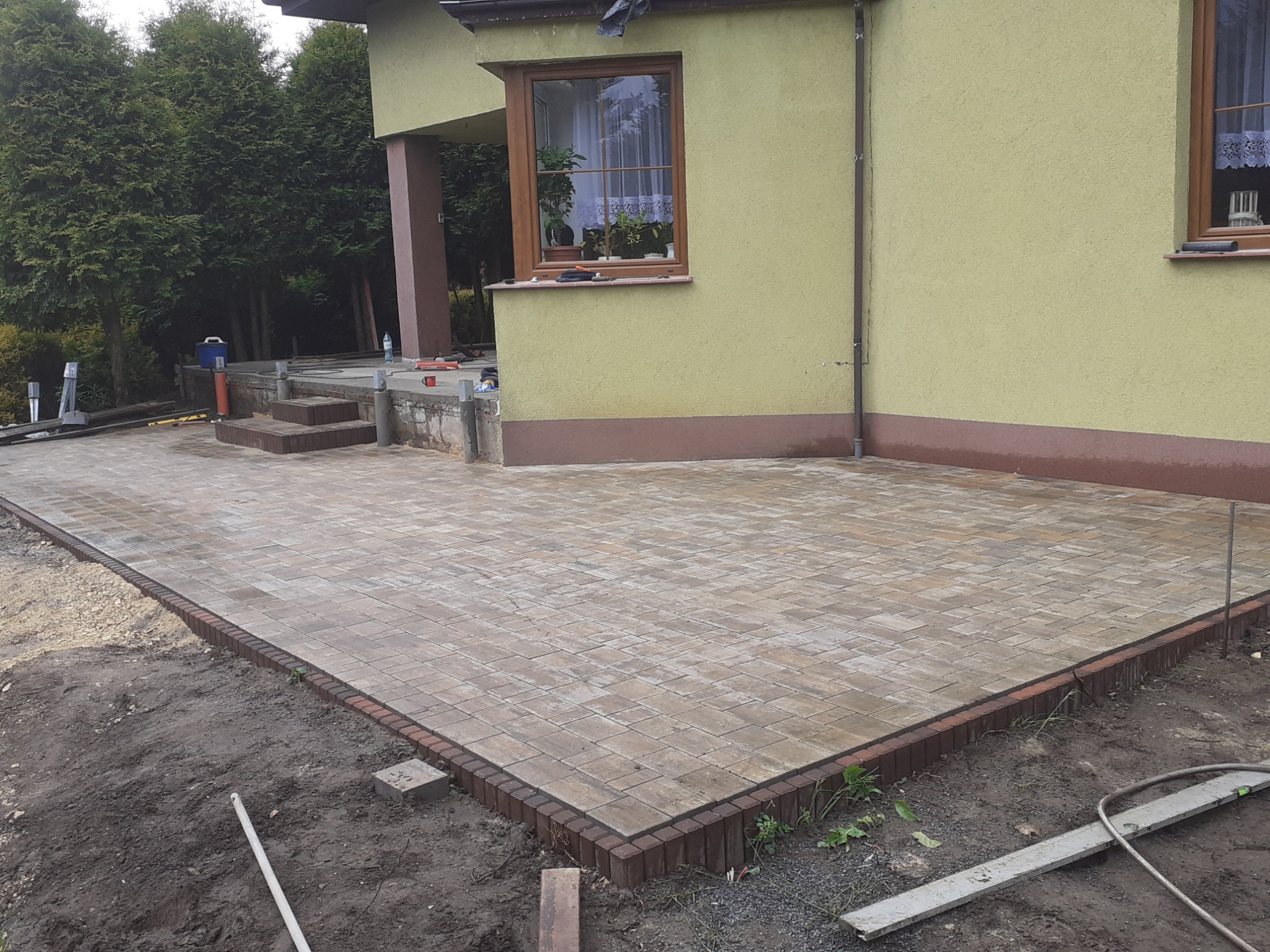 Ułożona kostka brukowa w odcieniach beżu i brązu przed domem z zieloną elewacją i brązowymi detalami, widoczne narzędzia i elementy przygotowania terenu.
