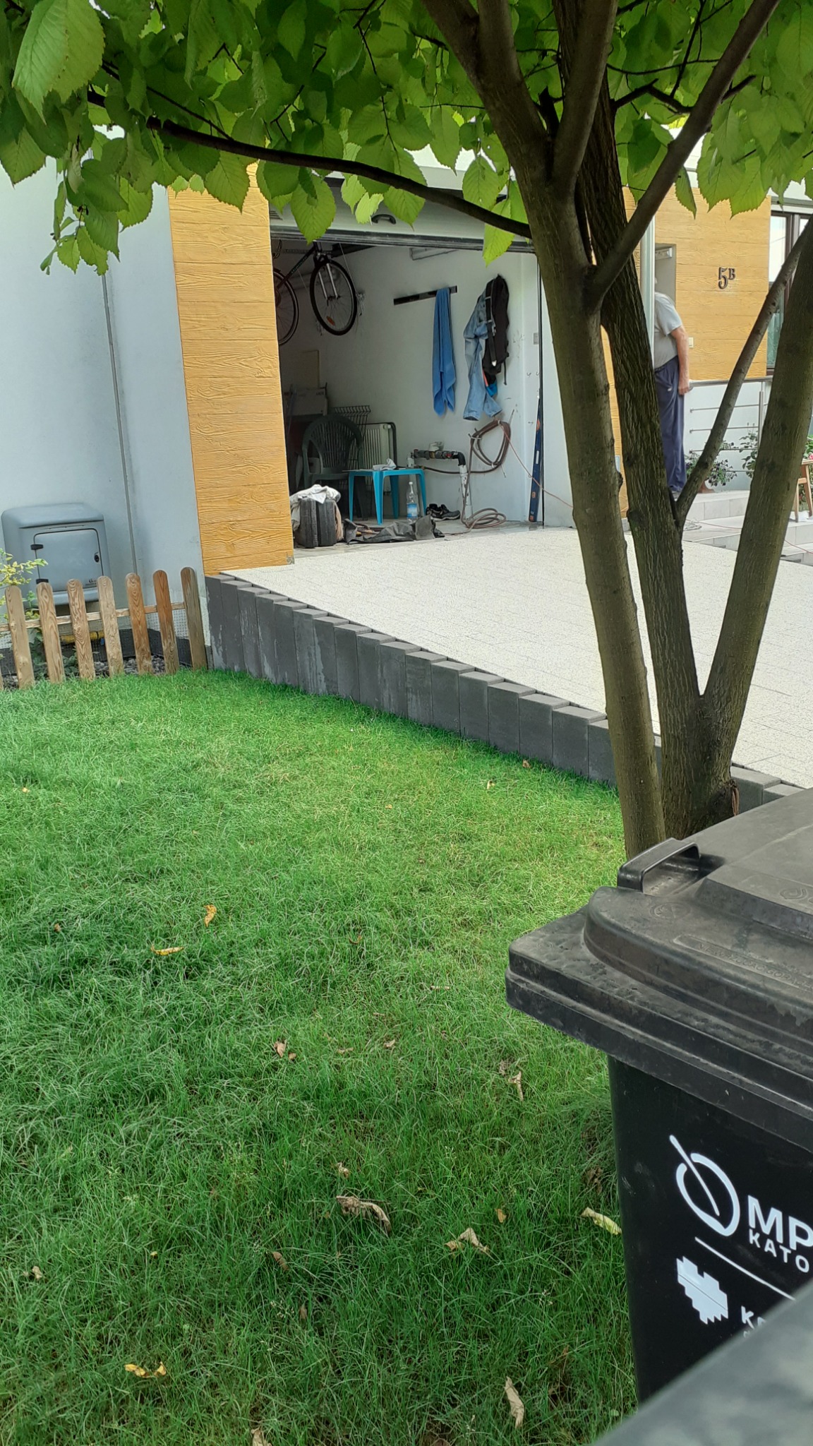 Szara kostka obrzeżowa oddzielająca trawnik od podjazdu z żywicy epoksydowej, widok na otwarty garaż z rowerem i narzędziami.