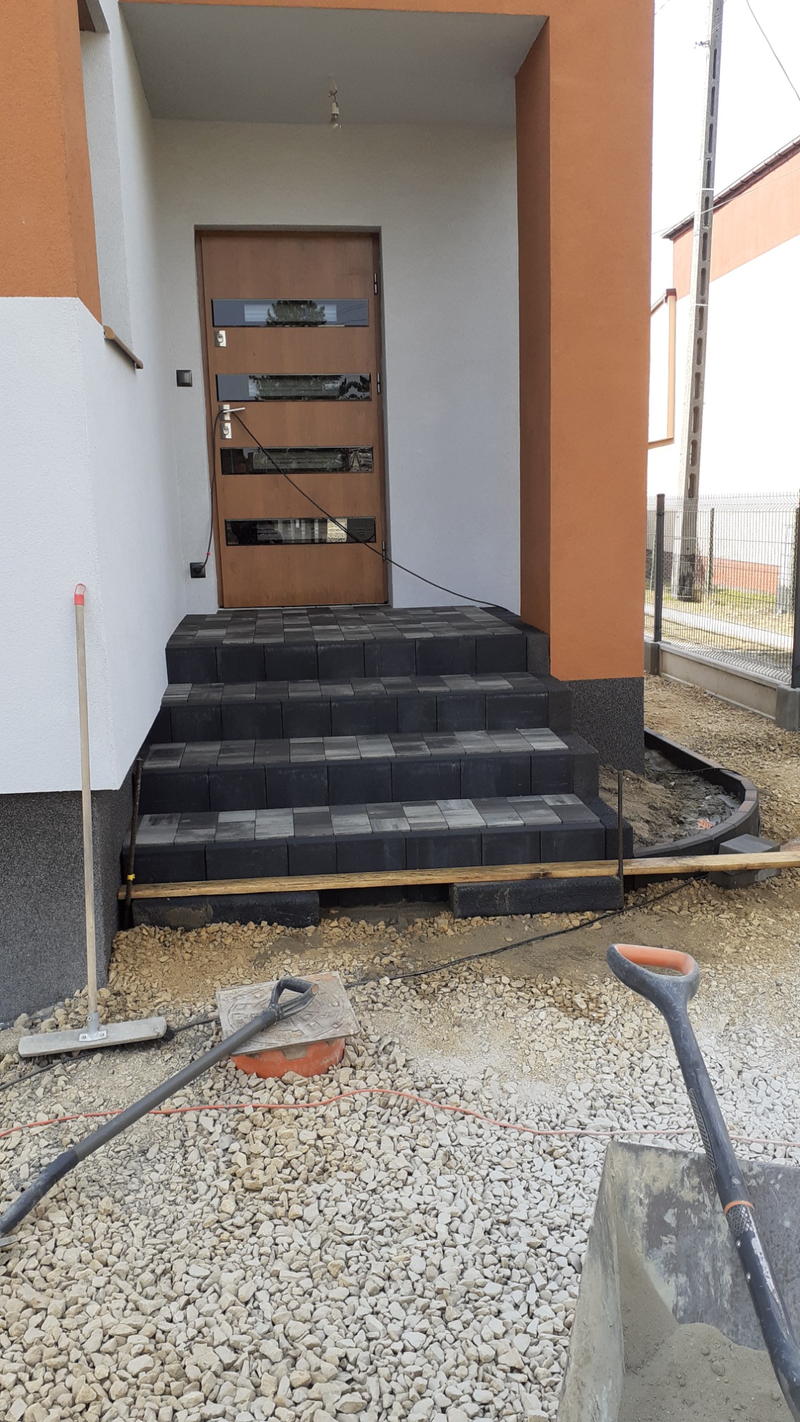Schody wejściowe do domu wyłożone czarno-szarą kostką brukową, widoczne narzędzia brukarskie i przygotowane podłoże z kruszywa.