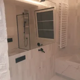 Wykończona łazienka z białymi płytkami imitującymi marmur, czarną armaturą i kabiną prysznicową z czarnymi profilami, widoczna w lustrze.