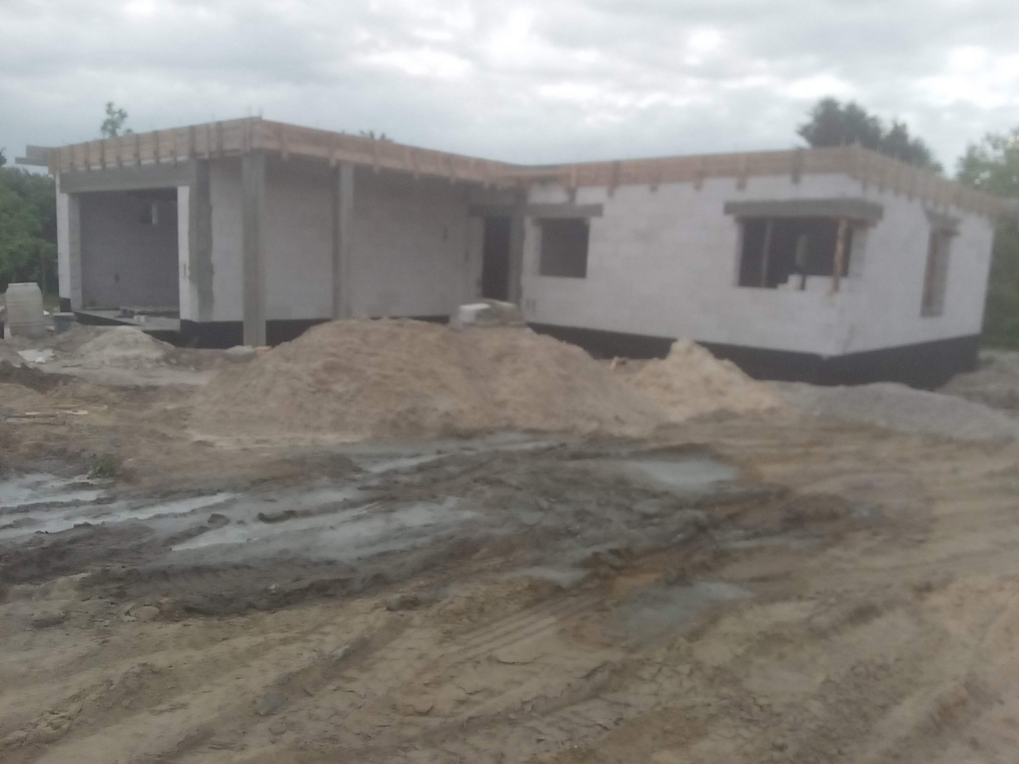 Stan surowy otwarty domu z garażem w trakcie budowy, widoczne szalunki i materiały budowlane na błotnistym terenie.