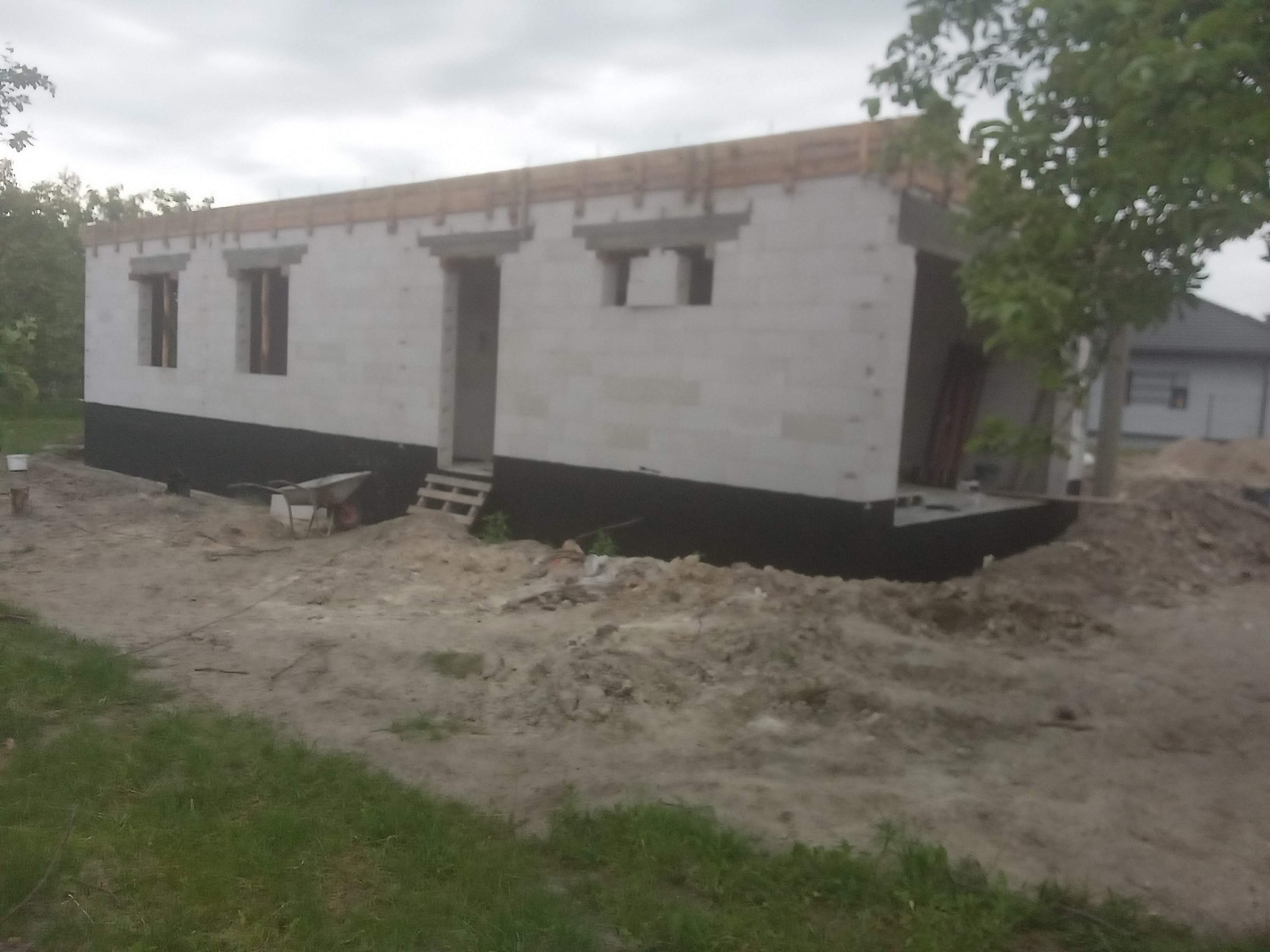 Budowa domu z bloczków betonowych, widoczne okna i drzwi w stanie surowym, teren budowy z ziemią i drzewami w tle. Fundament zabezpieczony hydroizolacją.
