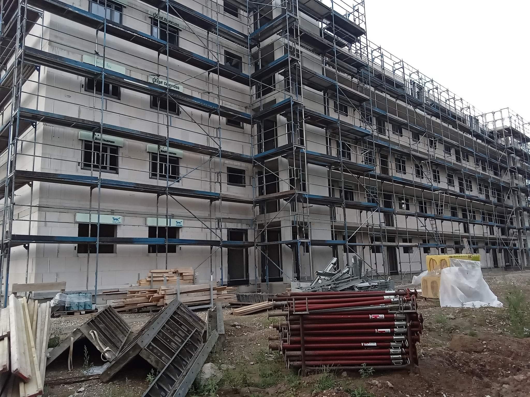 Wielopiętrowy budynek w trakcie budowy, otoczony rusztowaniami. Widoczne materiały budowlane i elementy konstrukcyjne. Budynek z białego bloczka.