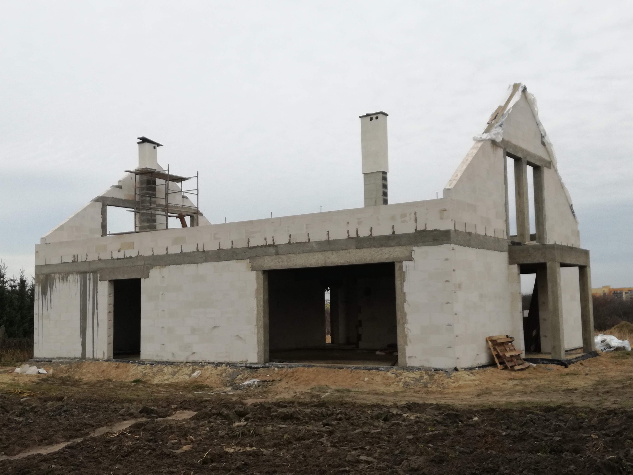 Surowy stan domu z białych pustaków, widoczne otwory okienne i drzwiowe, komin. Na dachu rusztowanie. Teren wokół domu z ziemią i resztkami materiałów budowlanych.