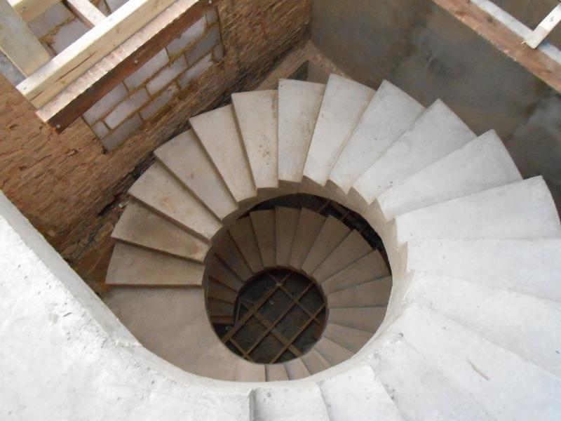 Spiralne schody z betonu w surowym stanie, widok z góry na okrągły otwór. Mur z cegły i drewniane elementy konstrukcyjne w tle.
