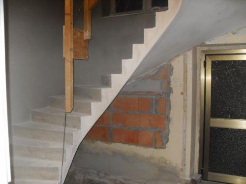 Spiralne, surowe schody z betonu w trakcie budowy, widoczne cegły i drewniane podpory. Ujęcie z dołu, z widoczną częścią drzwi po prawej stronie.