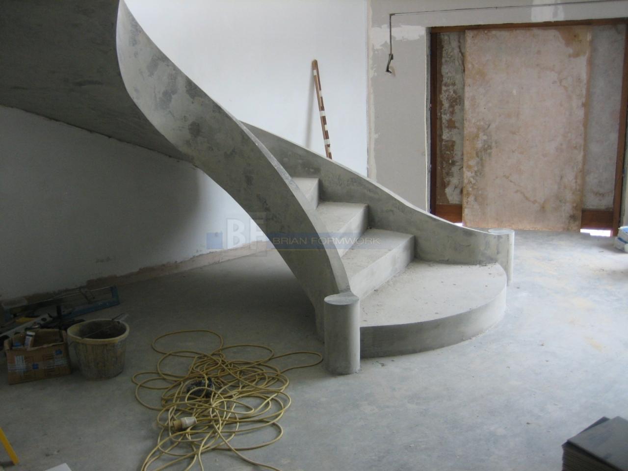 Spiralne schody betonowe w surowym stanie, z widocznymi elementami szalunku i niedokończonym wykończeniem wnętrza, w tle otwarte drzwi i drabina.