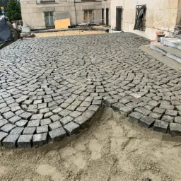 Fragment układanej nawierzchni z szarej kostki granitowej w kształcie wachlarza na podsypce piaskowej przed budynkiem.