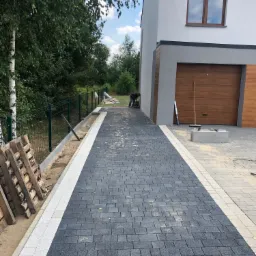 Nowo ułożona, ciemnoszara kostka na podjeździe do domu z brązową bramą garażową, obramowana jasną kostką. Widoczny pracownik zagęszczający podłoże, paleta z kostką i przygotowane obrzeża.