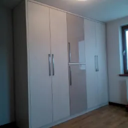 Duża, jasna szafa wnękowa z frontami w odcieniach bieli i szarości, z długimi, pionowymi uchwytami, dopasowana do wysokości pomieszczenia, na drewnianej podłodze.
