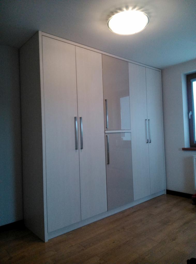 Duża, jasna szafa wnękowa z frontami w odcieniach bieli i szarości, z długimi, pionowymi uchwytami, dopasowana do wysokości pomieszczenia, na drewnianej podłodze.