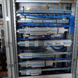 Rozdzielnica elektryczna z precyzyjnie ułożonymi niebieskimi przewodami, widoczne zabezpieczenia i szyny DIN. Estetyczny montaż w metalowej obudowie.