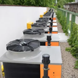 Szereg zainstalowanych, pomarańczowo-szarych urządzeń grzewczych z wentylatorami, ustawionych wzdłuż białego muru i ogrodzenia z zieloną roślinnością, na żwirowym podłożu.