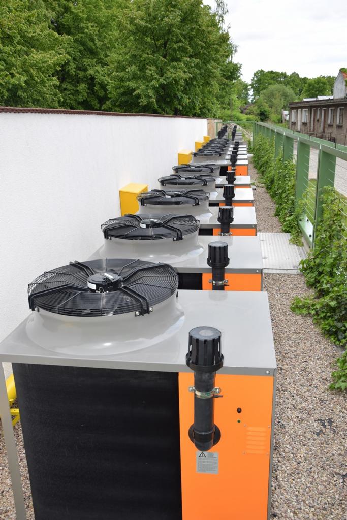 Szereg zainstalowanych, pomarańczowo-szarych urządzeń grzewczych z wentylatorami, ustawionych wzdłuż białego muru i ogrodzenia z zieloną roślinnością, na żwirowym podłożu.