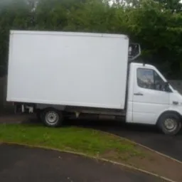 Biała furgonetka chłodnia zaparkowana na poboczu, widok z boku, z zielonymi drzewami w tle. Widoczny agregat chłodniczy.