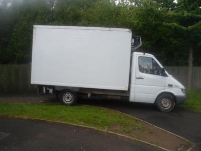 Biała furgonetka chłodnia zaparkowana na poboczu, widok z boku, z zielonymi drzewami w tle. Widoczny agregat chłodniczy.