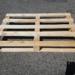 Jasna, drewniana paleta transportowa, widoczna z góry na szarym, betonowym podłożu w słoneczny dzień. Widoczne ślady użytkowania.
