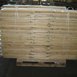 Stos desek z metalowymi wzmocnieniami na palecie EPAL, spięty sznurkiem. Widoczne niedoskonałości drewna i faktura betonu na podłodze.