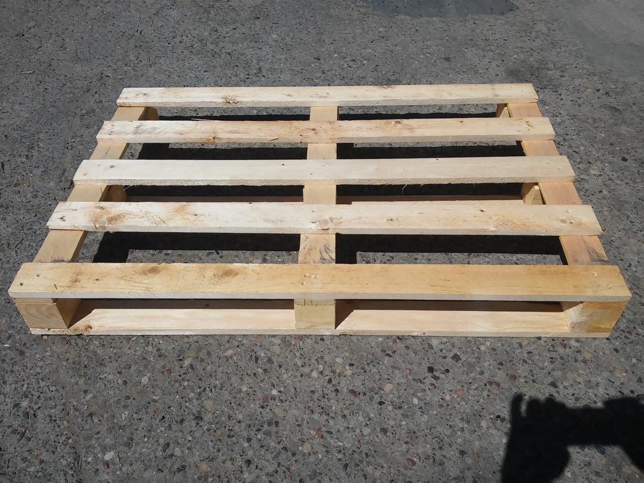 Jasna, drewniana paleta transportowa, widoczna z góry na szarym, betonowym podłożu w słoneczny dzień. Widoczne ślady użytkowania.