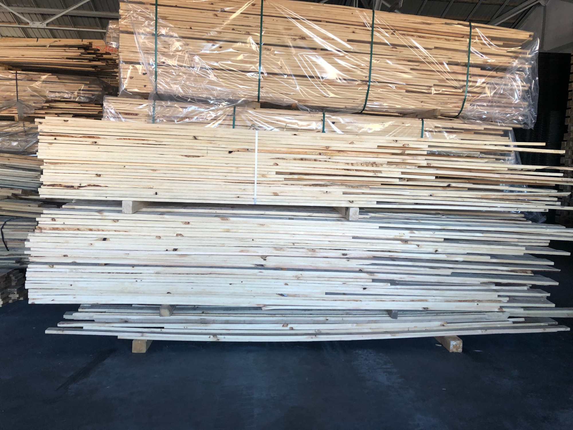 Stosy jasnego, surowego drewna ułożone na paletach w magazynie, część zafoliowana, widoczne sęki i naturalne przebarwienia drewna.