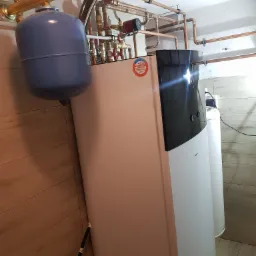 Kotłownia z nowoczesnym piecem dwufunkcyjnym Daikin, instalacją z miedzianych rur i zbiornikiem wyrównawczym z manometrem, na tle paneli ściennych imitujących drewno.