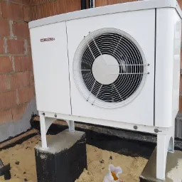 Biała pompa ciepła marki Stiebel Eltron zamontowana na betonowych fundamentach przy ścianie z cegły dziurawki, widoczna łopata i piasek.