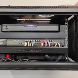 Czarna szafa rack z zamontowanymi urządzeniami sieciowymi: rejestrator, switch TP-Link z podłączonymi kablami Ethernet w różnych kolorach, listwa zasilająca StalFlex, widoczny wentylator chłodzący.
