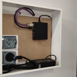 Wirekom Piotr Dworzyński - Wnętrze białej szafki z osprzętem telekomunikacyjnym: czarny router przymocowany do korkowej płyty, dwa gniazdka elektryczne z przezroczystymi klapkami, zasilacz i kable. Widoczne szare ściany.