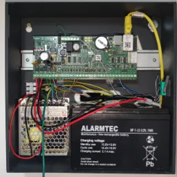 Wnętrze skrzynki alarmowej z płytą główną Roger, zasilaczem impulsowym i akumulatorem Alarmtec BP 7-12, podłączone kable i wtyczka Ethernet. Widoczne oznaczenia i specyfikacje techniczne urządzeń.