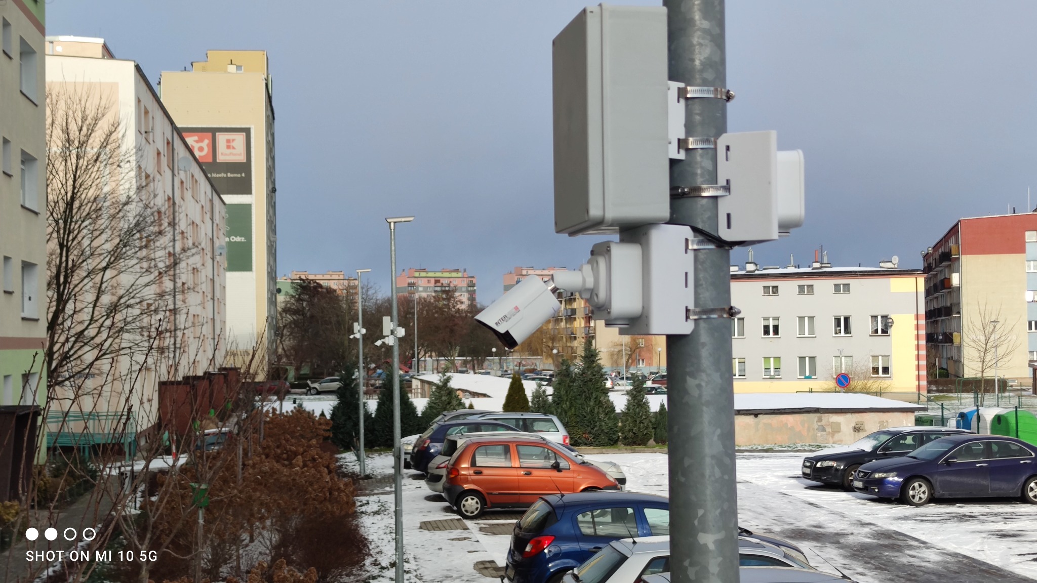 Zewnętrzna kamera monitoringu przymocowana do słupa na parkingu osiedlowym, z widocznymi puszkami instalacyjnymi i budynkami w tle, w zimowej scenerii.