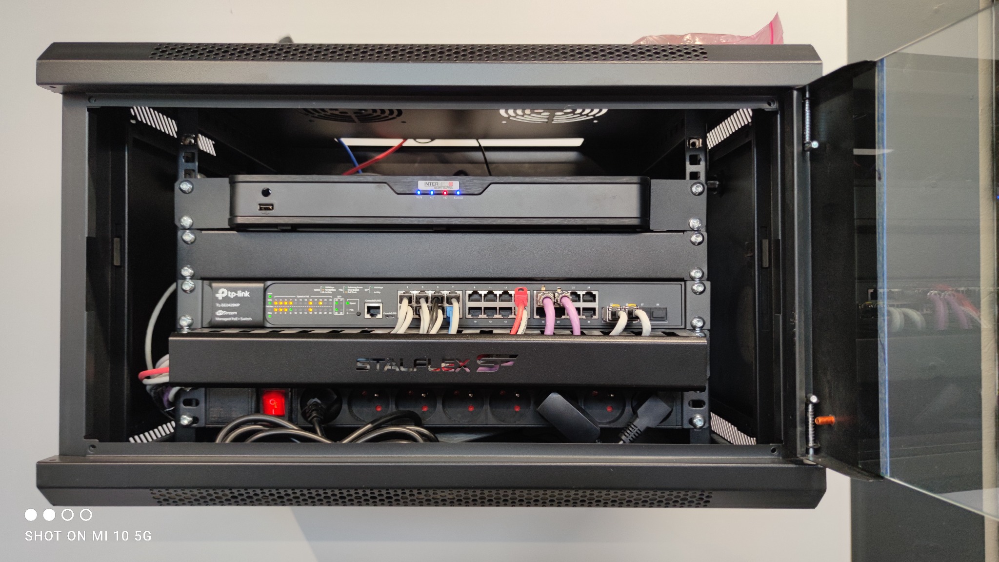 Czarna szafa rack z zamontowanymi urządzeniami sieciowymi: rejestrator, switch TP-Link z podłączonymi kablami Ethernet w różnych kolorach, listwa zasilająca StalFlex, widoczny wentylator chłodzący.