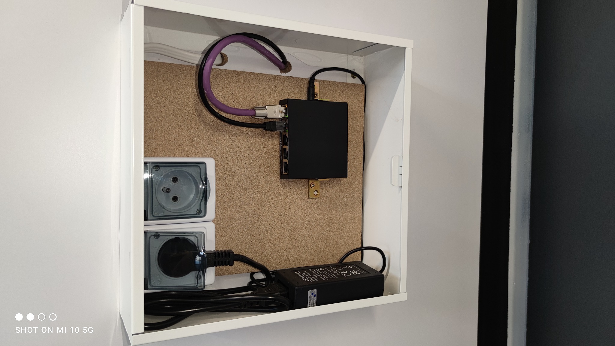 Wnętrze białej szafki z osprzętem telekomunikacyjnym: czarny router przymocowany do korkowej płyty, dwa gniazdka elektryczne z przezroczystymi klapkami, zasilacz i kable. Widoczne szare ściany.