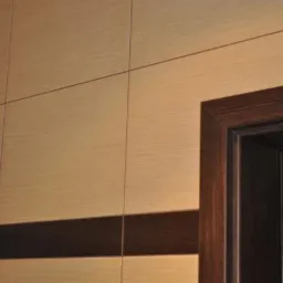 Fragment ściany wyłożonej jasnymi, drewnopodobnymi płytkami z ciemnobrązowym pasem dekoracyjnym i ciemnobrązową ościeżnicą drzwiową.