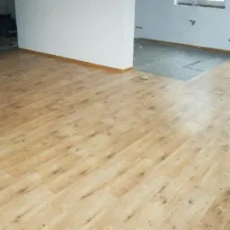 Jasne panele podłogowe imitujące drewno, ułożone w nowym pomieszczeniu z białymi ścianami, widoczny fragment podłogi w innym pokoju z szarą wylewką.