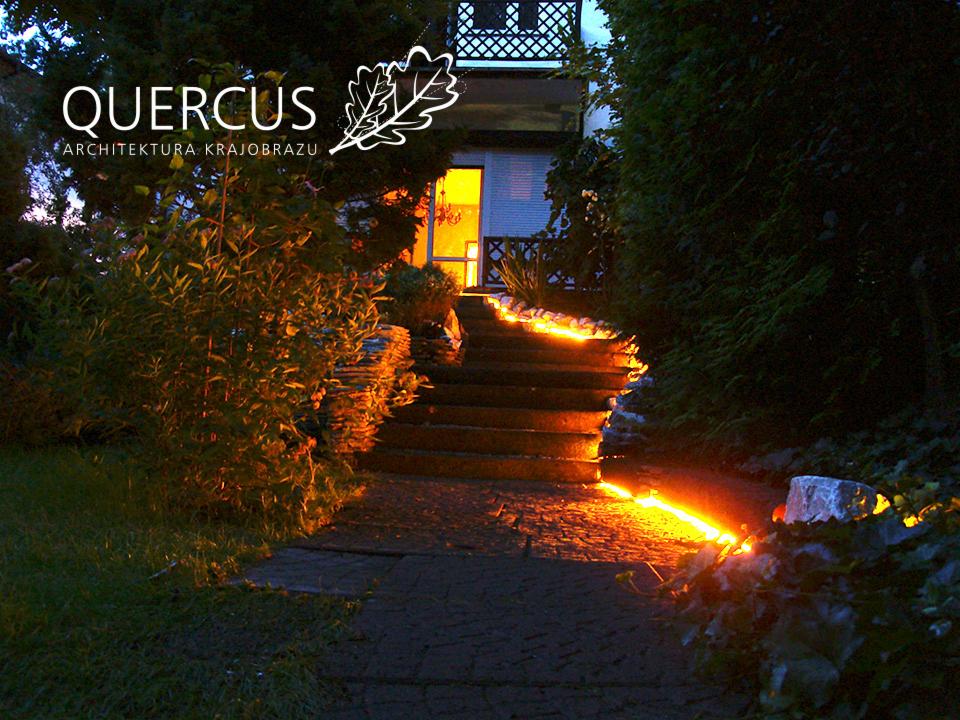 Oświetlona ścieżka z kamienia prowadzi do domu. Schody podkreślone ciepłym światłem, logo firmy architektonicznej w lewym górnym rogu. Nocne ujęcie.