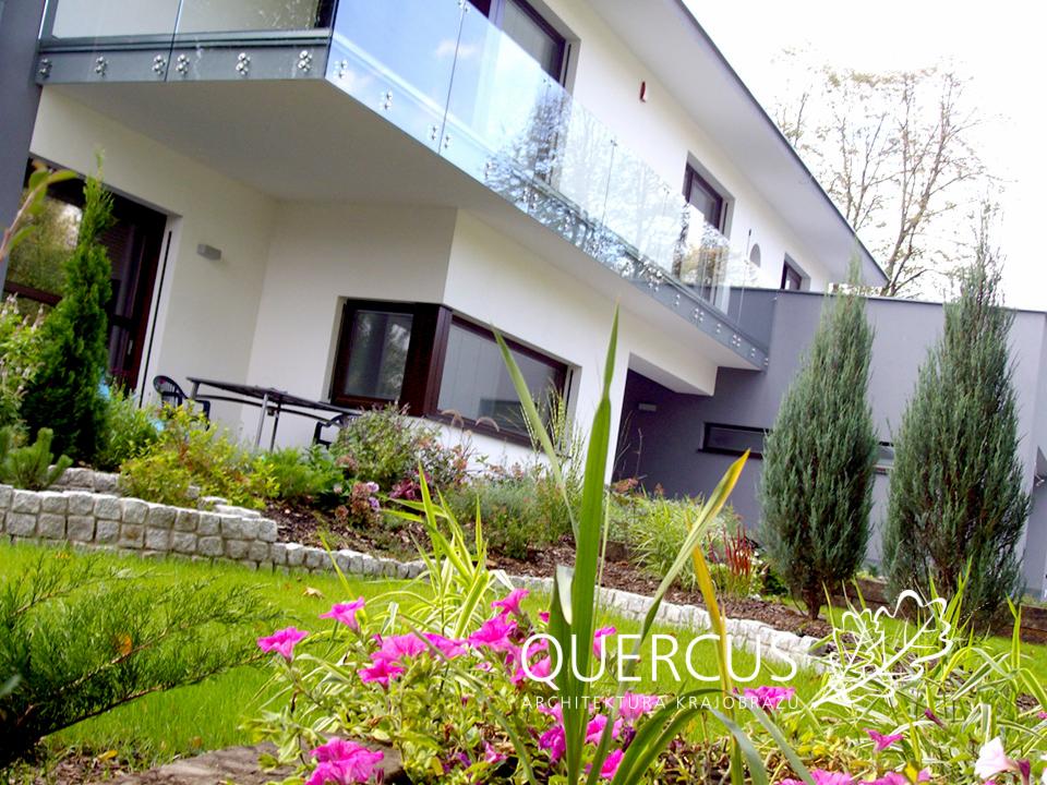 Nowoczesny ogród z kwitnącymi różowymi kwiatami na pierwszym planie, elegancki dom z balkonem i szklaną balustradą w tle. Granitowa obudowa rabat.