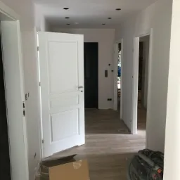 Korytarz po remoncie z widocznymi trzema parami białych drzwi wewnętrznych, parkietem, punktowym oświetleniem sufitowym oraz kartonem i odkurzaczem przemysłowym w trakcie sprzątania.