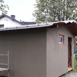 Świeżo otynkowany budynek gospodarczy z małym oknem, widoczny pracownik przygotowujący zaprawę, w tle domy jednorodzinne i zieleń.