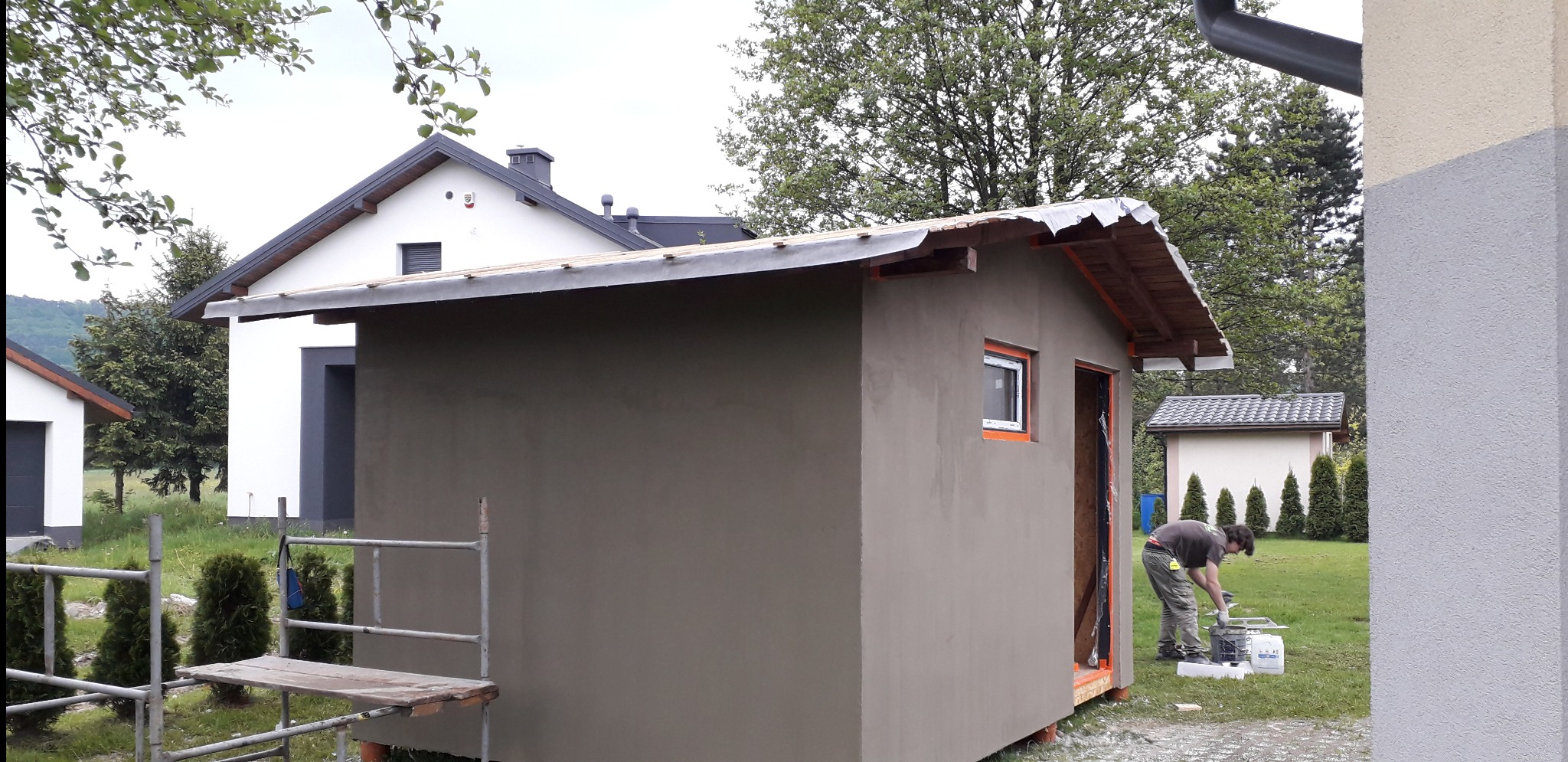 Świeżo otynkowany budynek gospodarczy z małym oknem, widoczny pracownik przygotowujący zaprawę, w tle domy jednorodzinne i zieleń.