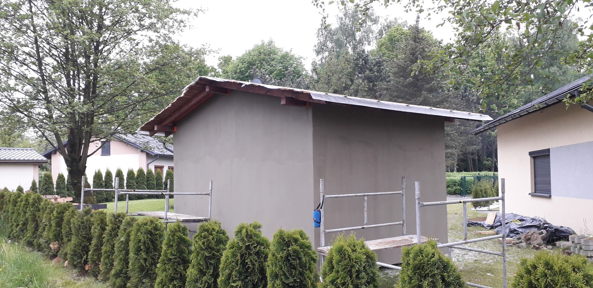 Budynek gospodarczy w trakcie tynkowania, widoczne rusztowania i świeżo nałożony tynk w kolorze szarym, w tle inne budynki i drzewa.
